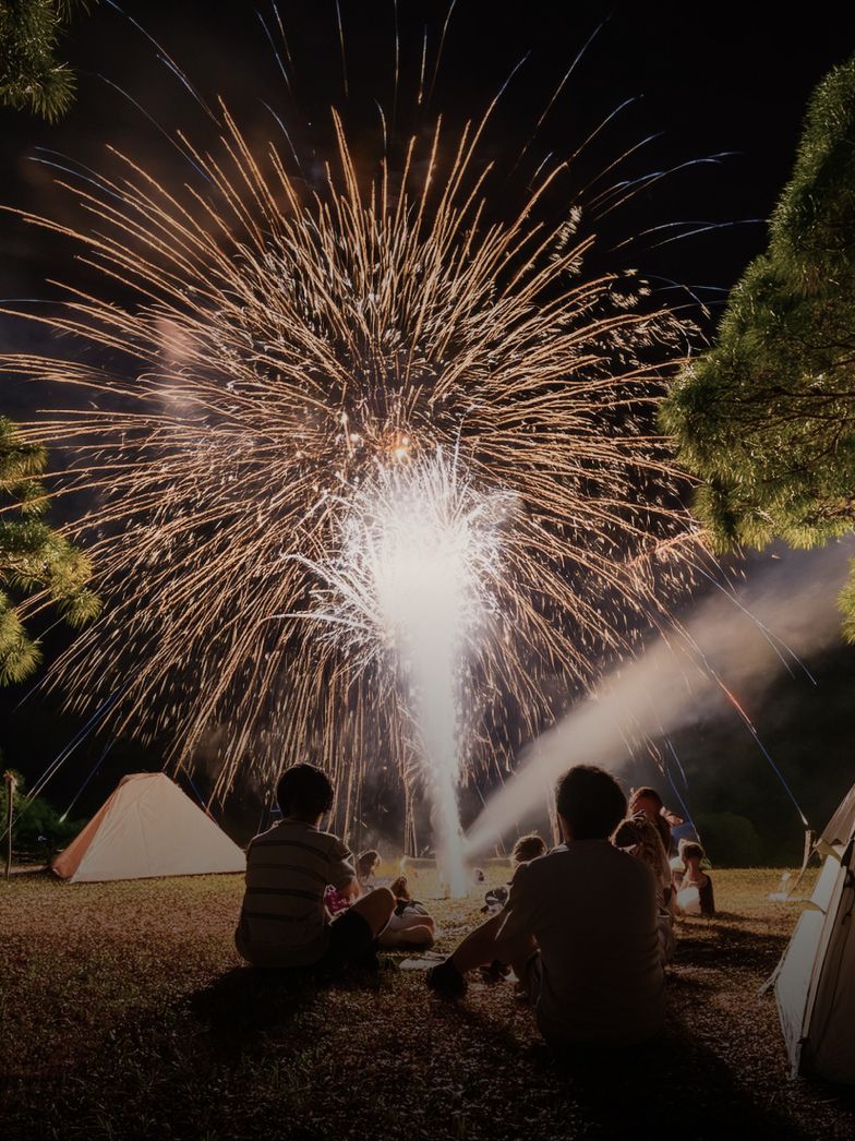 夜のキャンプ場で、テントの近くに座る複数のキャンパーたちが、置き型の花火を見て楽しんでいる